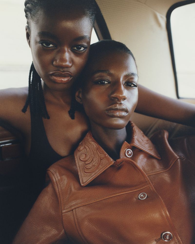 Two young African-descent women in a car wearing a brown leather jacket and a black top, gazing directly at the camera with a serious expression.