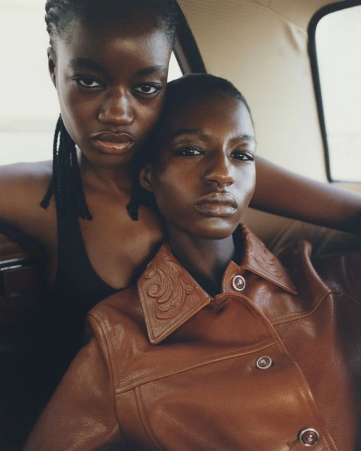 Two young African-descent women in a car wearing a brown leather jacket and a black top, gazing directly at the camera with a serious expression.