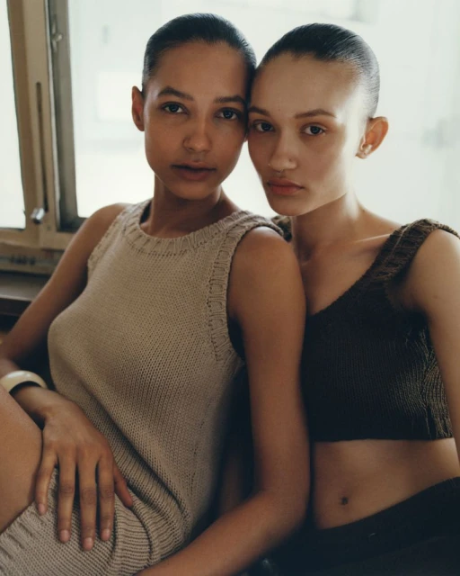 Two women pose indoors, showcasing knitwear fashion. One wears a beige dress, while the other dons a brown crop top and dark pants.