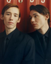 Two men in dark suits and ties posing closely together in front of a vertical red corrugated metal wall.