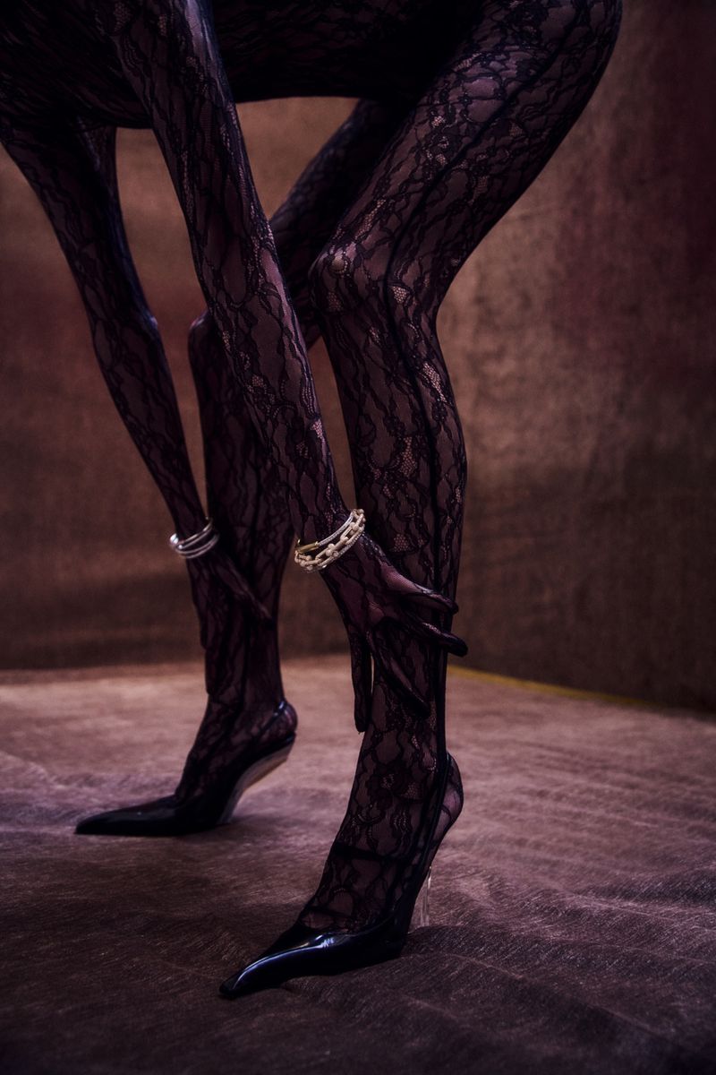 Close-up studio shot of a person wearing black lace hosiery and black patent leather heels, accessorized with silver and gold anklets.