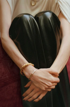 Seated woman in leather pants rests arms on knees, wearing gold bracelets, illuminated by soft studio light against a neutral backdrop.