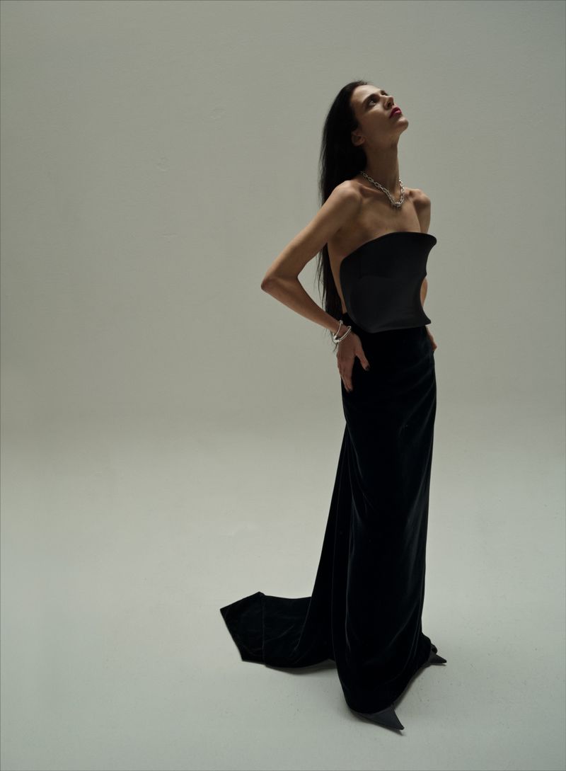 A slender, dark-haired woman in a strapless black gown gazes upwards in a studio setting with neutral background and diffused lighting.