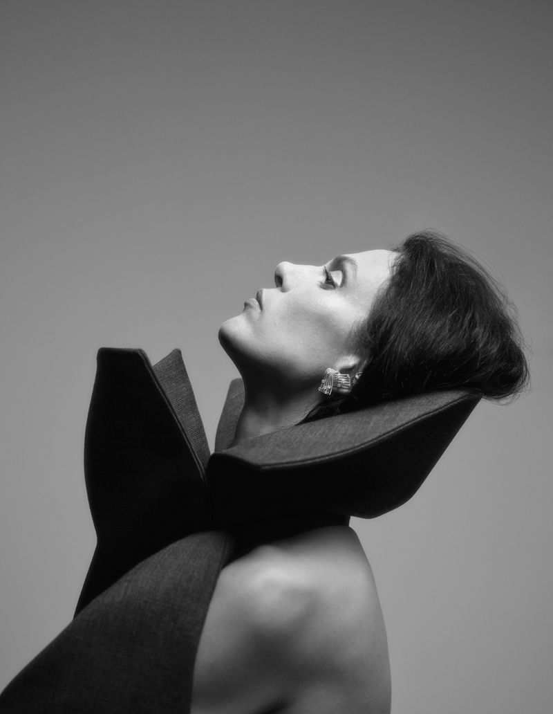 Black and white portrait of a woman with dark hair and jewelry, wearing a large collared garment, head tilted back against a plain backdrop.