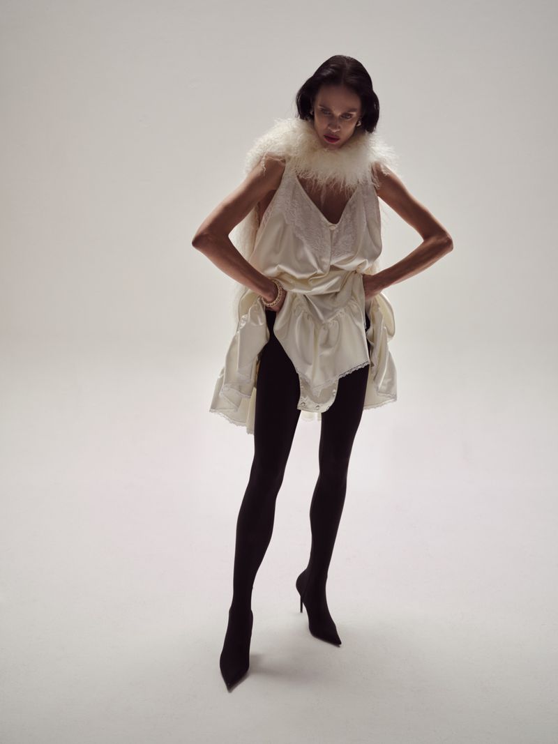 Full-length studio shot of a woman in white lingerie-inspired dress, black tights, and heels, against a bright neutral backdrop.
