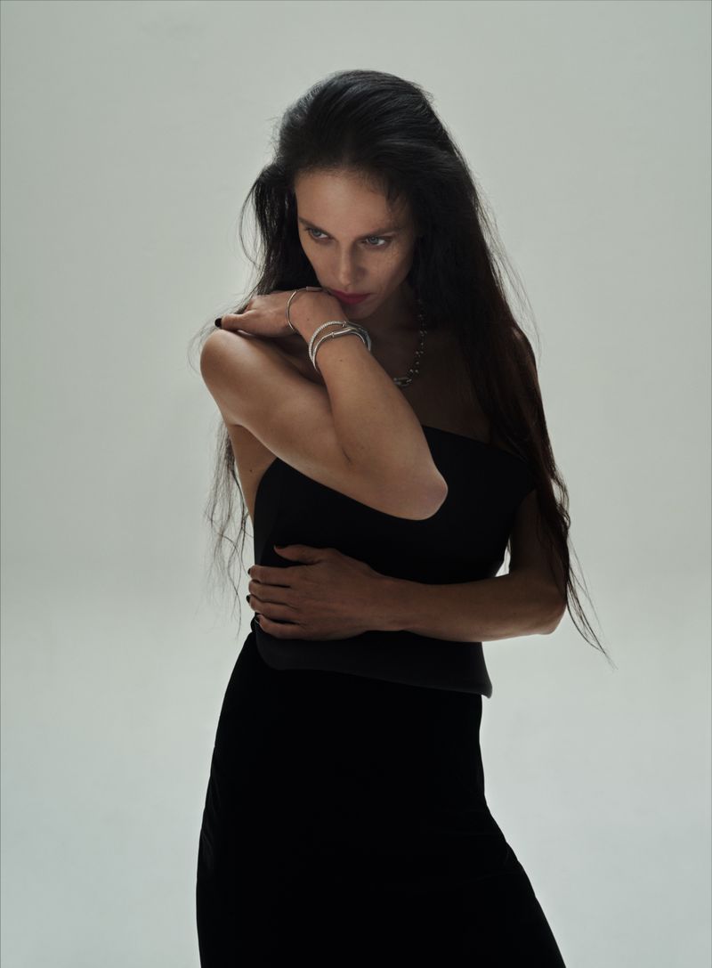 Model Aymeline Valade wearing a black dress and diamond jewelry against a light grey seamless studio background.