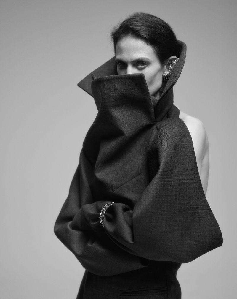 Monochrome fashion portrait of a woman in a sculptural garment with large collar and sleeves, posing against a neutral backdrop.