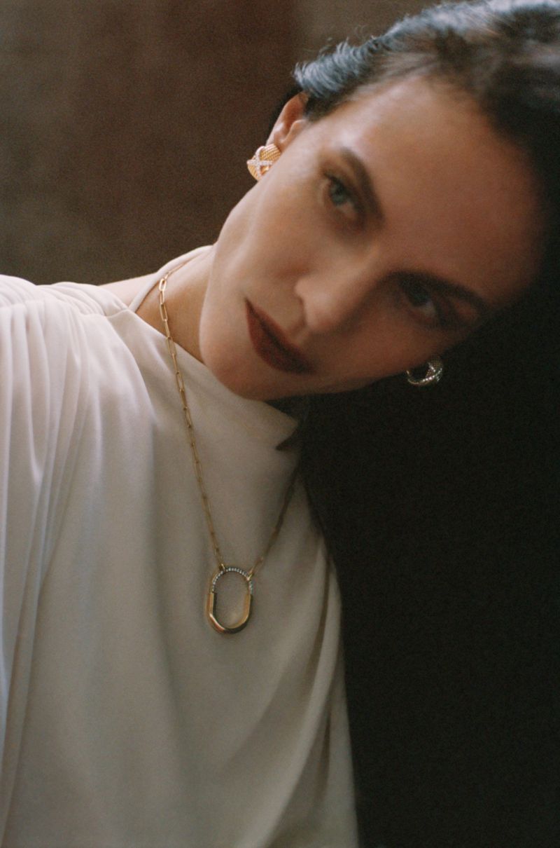 Close-up portrait of model Aymeline Valade wearing a white top and gold necklace against a dark textured background.