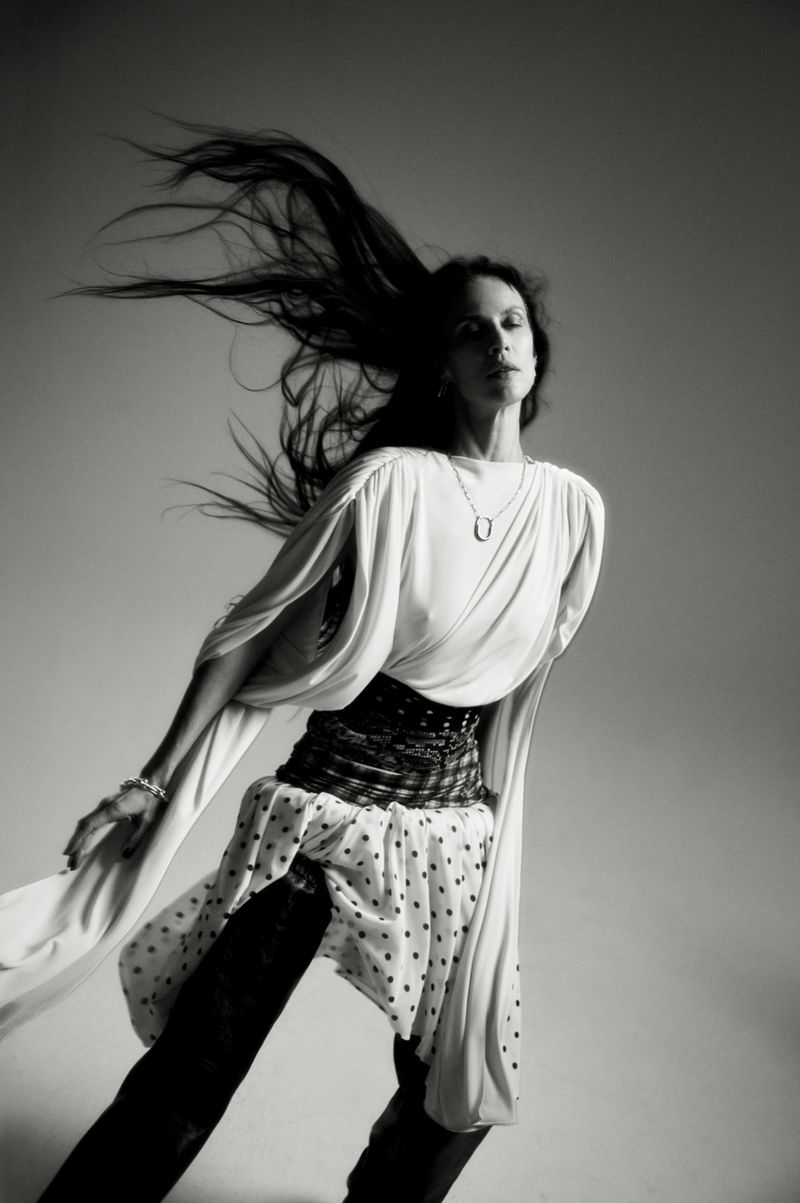 Model Aymeline Valade with long dark hair blowing in motion, wearing a white draped top and patterned skirt against a grey background.