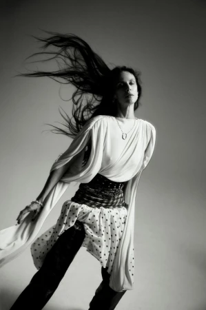 Model Aymeline Valade with long dark hair blowing in motion, wearing a white draped top and patterned skirt against a grey background.