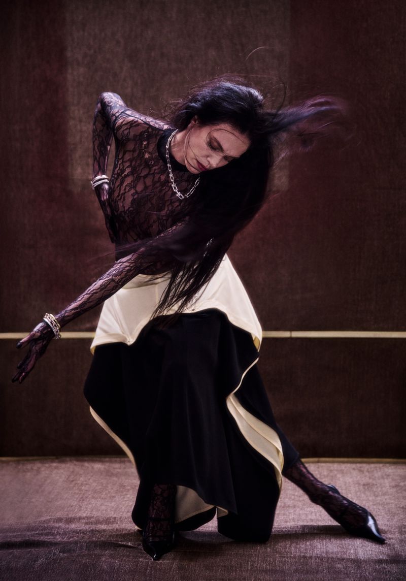 Female dancer in black lace top and flowing skirt poses expressively against a dark backdrop with dramatic lighting and shadows.