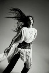 Model Aymeline Valade with long dark hair blowing in motion, wearing a white draped top and patterned skirt against a grey background.