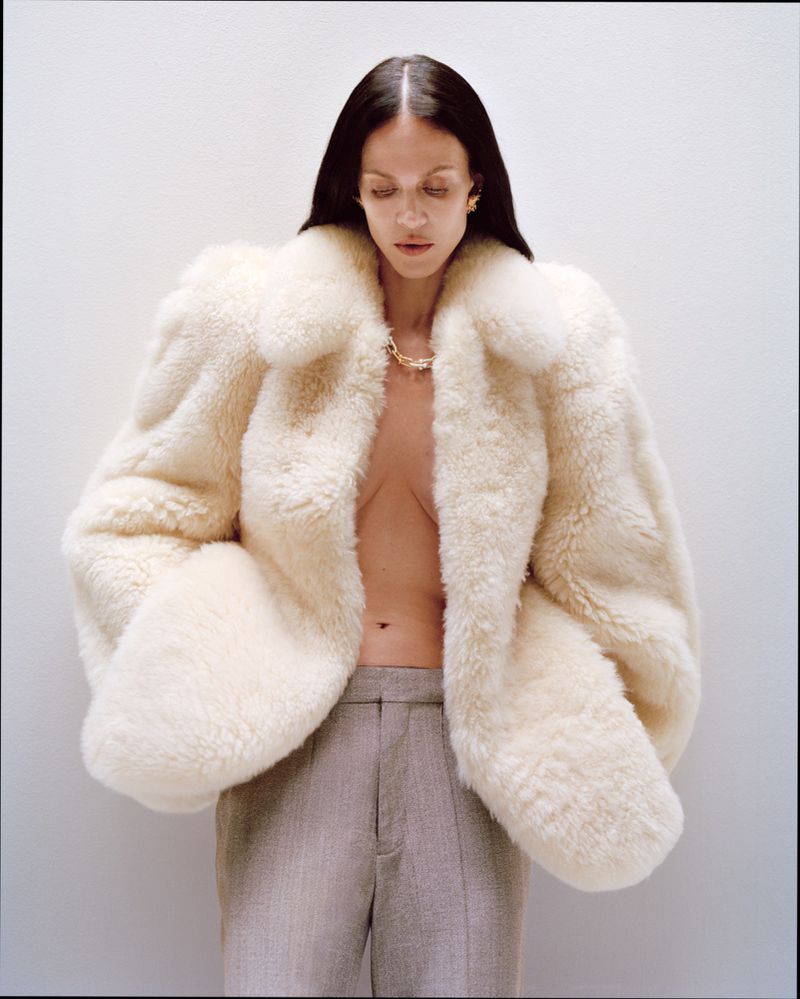 Model Aymeline Valade wearing a voluminous cream fur coat and grey trousers against a plain light background.