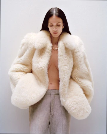 Model Aymeline Valade wearing a voluminous cream fur coat and grey trousers against a plain light background.