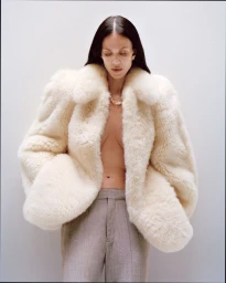 Model Aymeline Valade wearing a voluminous cream fur coat and grey trousers against a plain light background.