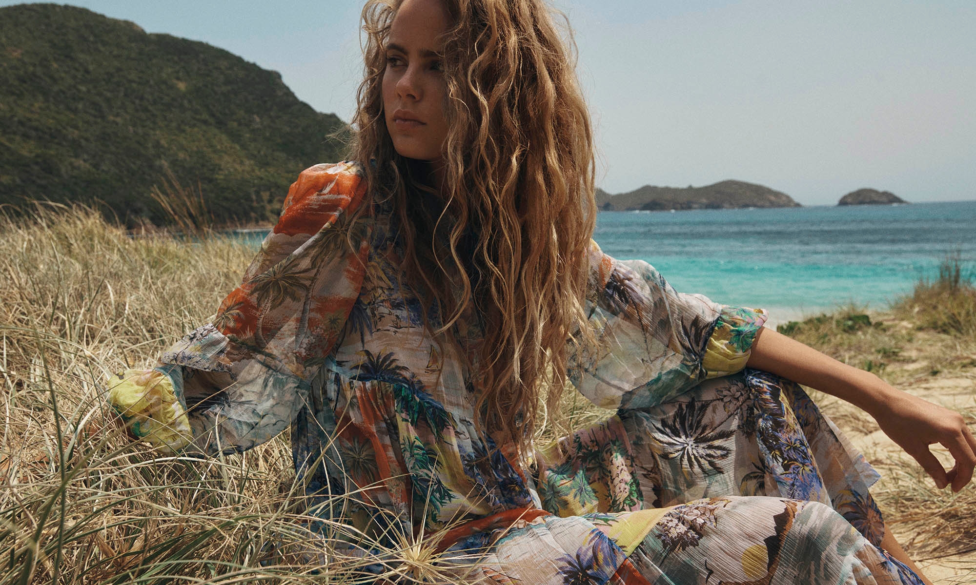 Adult woman sits in a grassy field near the beach, wearing a tropical print dress, with rolling hills and turquoise water in the background.