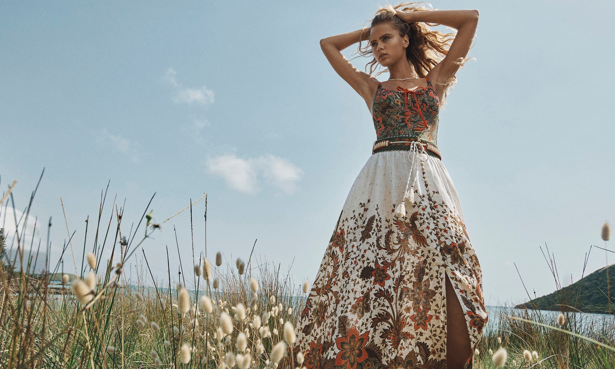 A woman in a floral dress stands in a grassy field under a bright blue sky, posing with her hands behind her head in natural light.