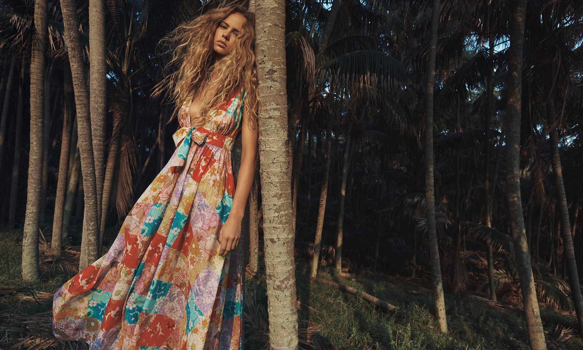 Adult female fashion model with long blonde hair wearing a floral patchwork sundress leaning against a palm tree in an outdoor jungle setting.