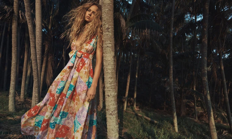 Adult female fashion model with long blonde hair wearing a floral patchwork sundress leaning against a palm tree in an outdoor jungle setting.