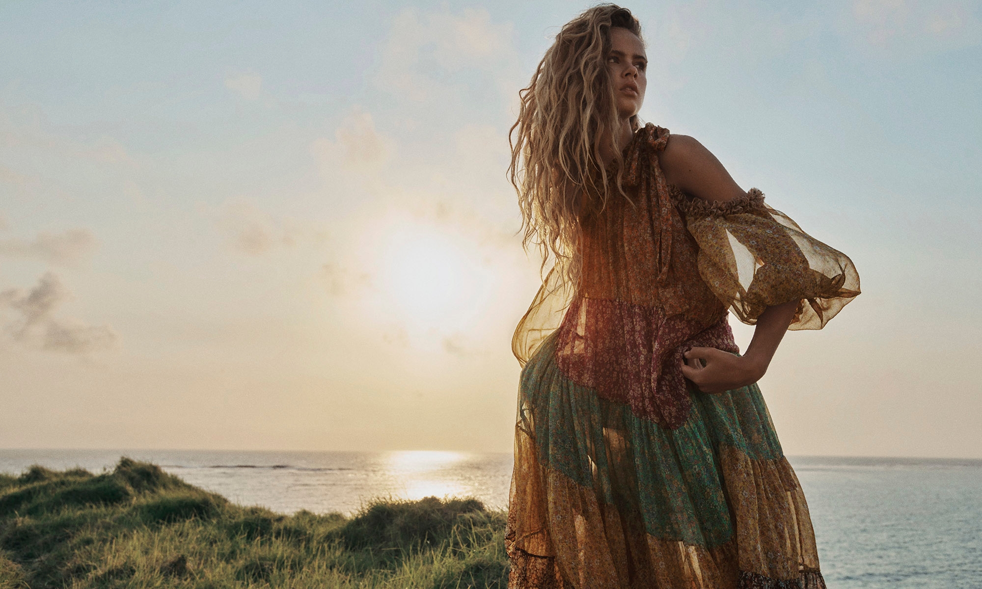 Woman with blonde hair posing in a flowing dress on a grassy hill overlooking the ocean at sunset in a fashion editorial shot.