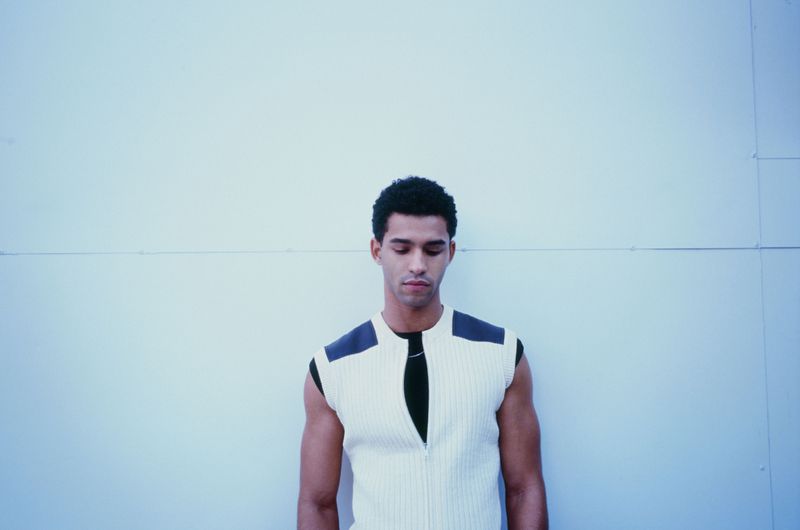 Young adult male in a sleeveless ribbed white shirt with black accents, stands indoors against a pale blue wall, eyes looking downward.