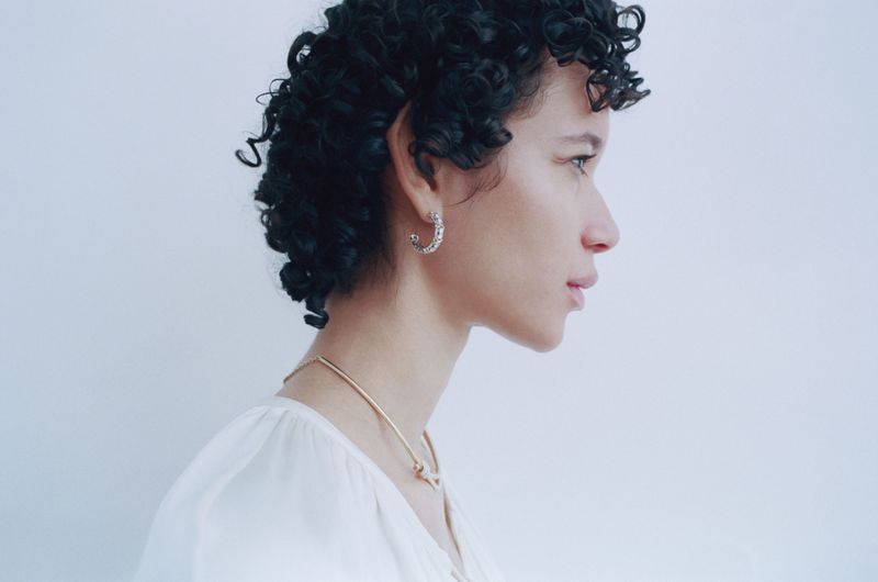 Close-up profile of a woman with curly hair wearing jewelry and a white blouse against a soft white background in a studio setting.