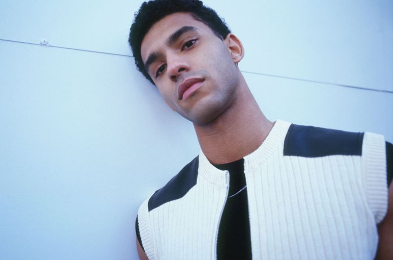 Close-up of a young adult male leaning against a wall wearing a sleeveless, cream-colored knit top with black panels on the shoulders.