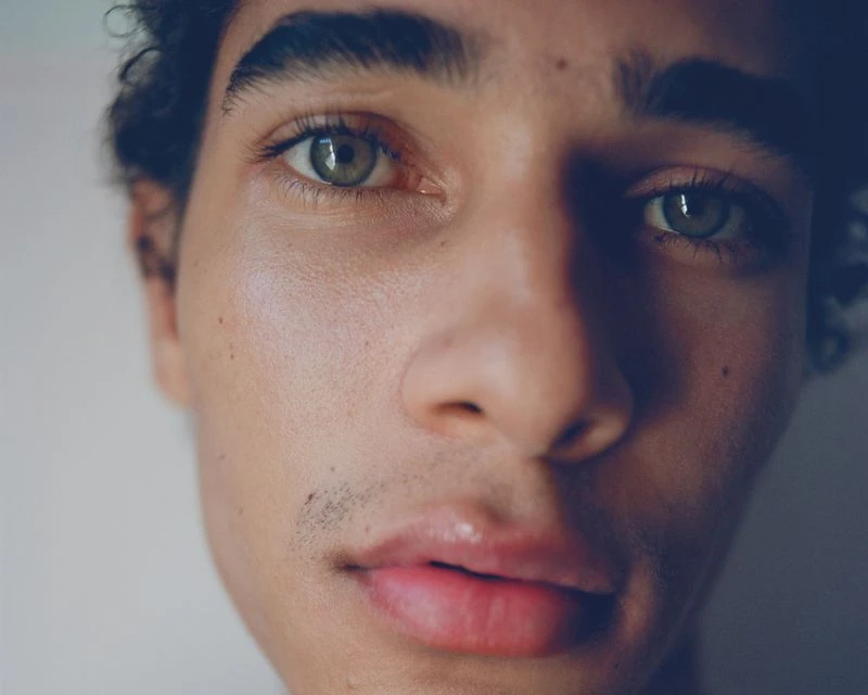 Close-up portrait of a young adult male with olive skin, green eyes, and dark curly hair against a neutral background.