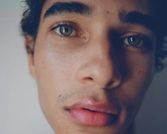 Close-up portrait of a young adult male with olive skin, green eyes, and dark curly hair against a neutral background.