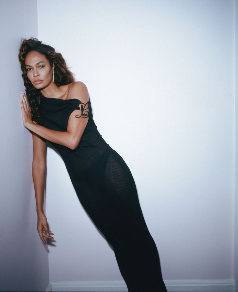 Elegant mixed-race woman in a black dress poses indoors, leaning against a pale corner wall. Fashion photography portrait.