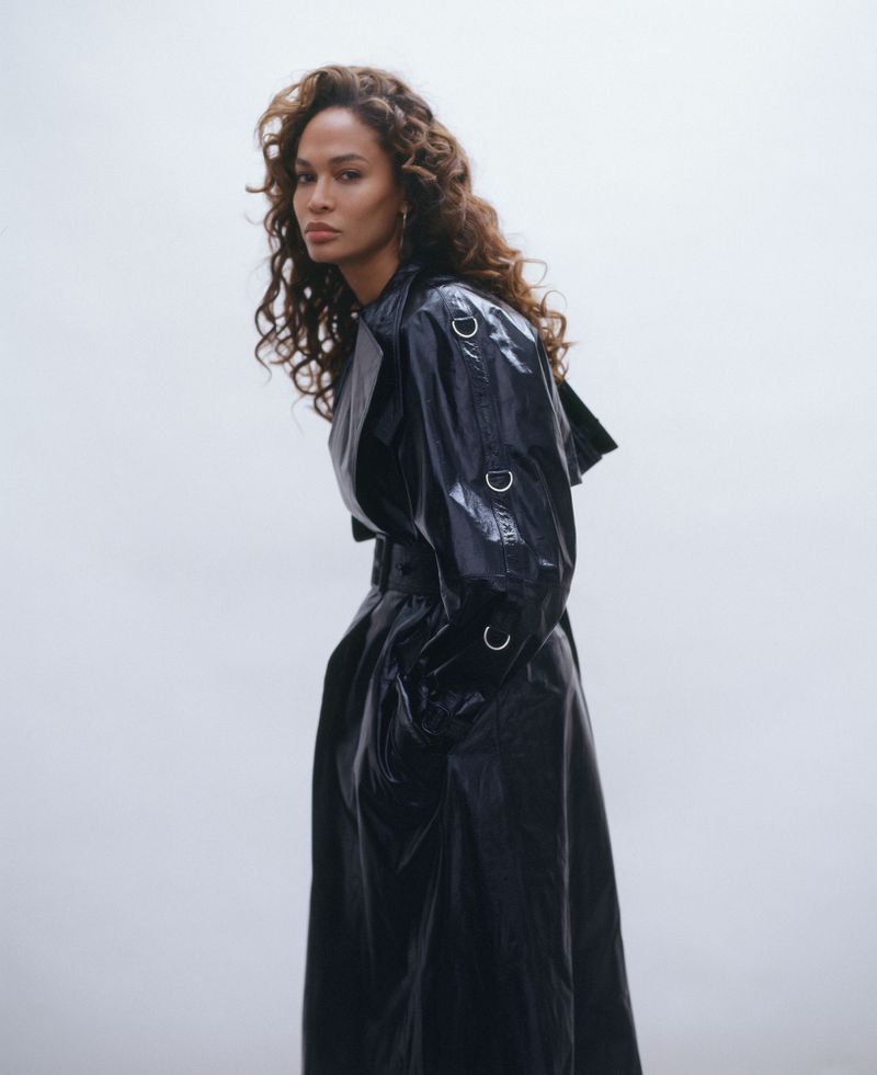 A woman with long brown hair poses in a glossy black trench coat against a soft white background, showcasing a modern fashion look.