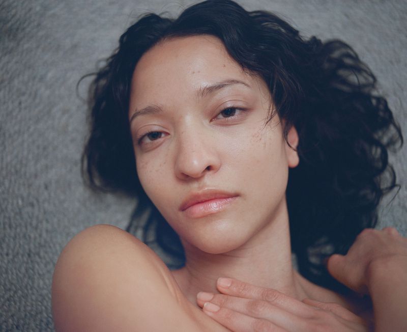 Close-up portrait of a young adult woman with dark, wavy hair, fair skin, and natural make-up laying against a gray textured background.