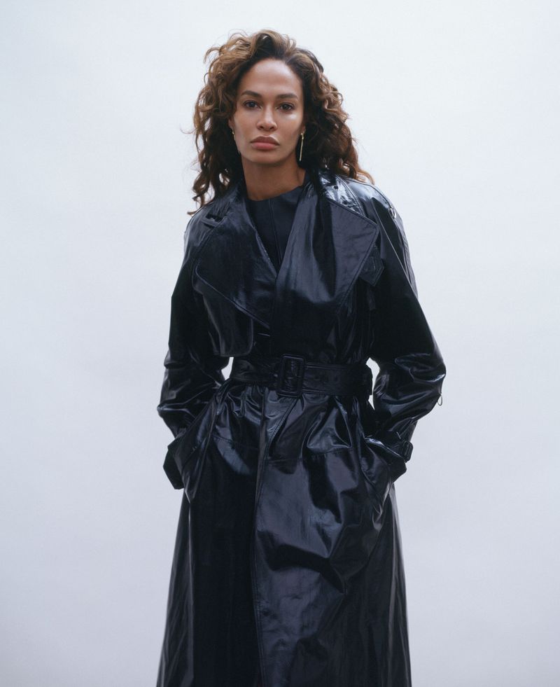 Portrait of an adult woman with curly brown hair wearing a shiny black trench coat looking at the camera against a light background.