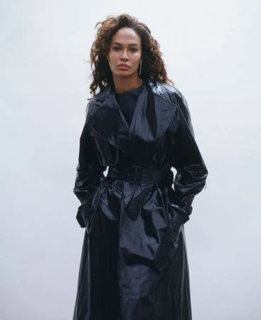Portrait of an adult woman with curly brown hair wearing a shiny black trench coat looking at the camera against a light background.