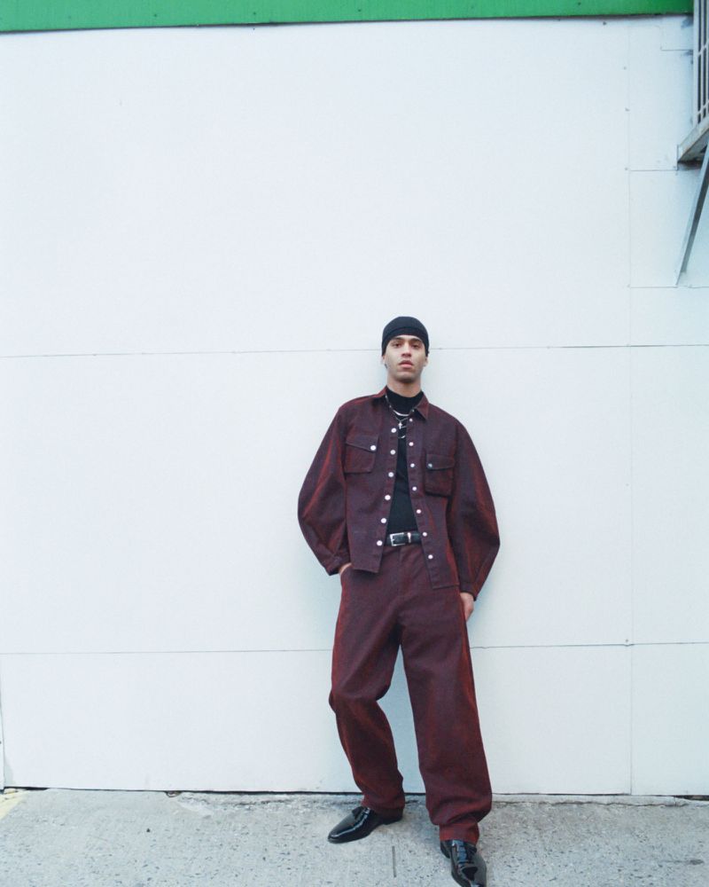 Male model in a maroon denim outfit stands against a white wall with green trim, showcasing a blend of casual and stylish fashion.