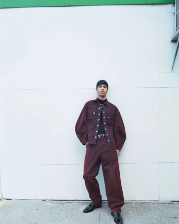 Male model in a maroon denim outfit stands against a white wall with green trim, showcasing a blend of casual and stylish fashion.