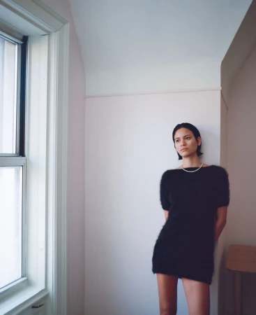 Model standing indoors wearing a black knit dress and pearl necklace next to a large window.