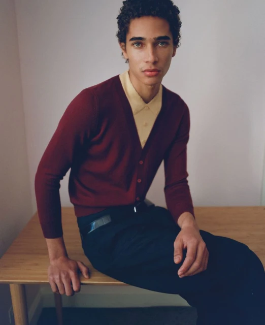 Portrait of a young adult male wearing a red cardigan and yellow collared shirt, seated on a wooden table in a studio setting.