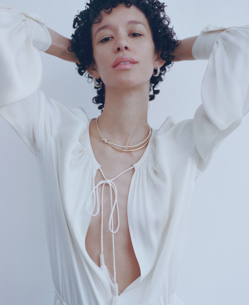 A fair-skinned young woman with curly hair poses in a white dress and gold necklace in a portrait against a light gray background.