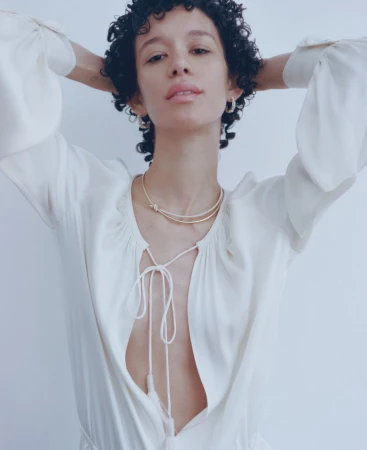 A fair-skinned young woman with curly hair poses in a white dress and gold necklace in a portrait against a light gray background.