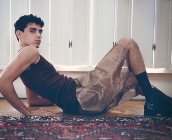 Male model poses indoors wearing a brown tank top and translucent shorts. He is laying on a red patterned carpet against a white wall.
