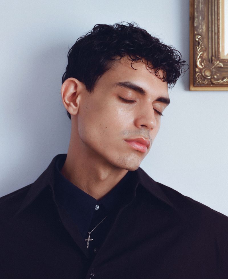 Portrait of a young adult man with closed eyes, wearing a black collared shirt and cross necklace, with an ornate picture frame in the background.