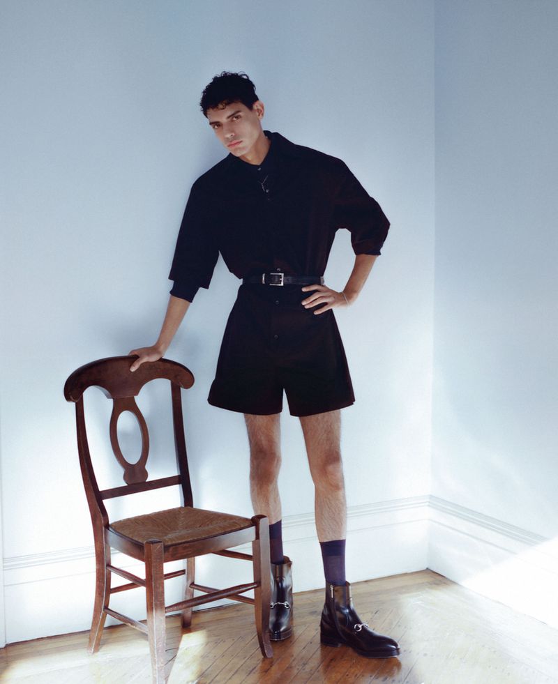 A young adult male in a black outfit poses near a wooden chair inside. He wears shorts, a button-down shirt, boots, and is leaning against a chair.