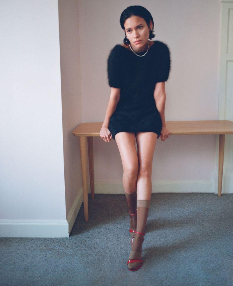 A woman in a black dress sitting on a table against a neutral background, wearing brown socks and red heels, posing for a fashion shot.