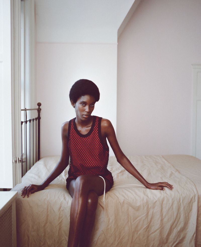 An adult woman with dark skin and a short afro hairstyle sits on a bed in a red mesh tank top and dark shorts in a bedroom.