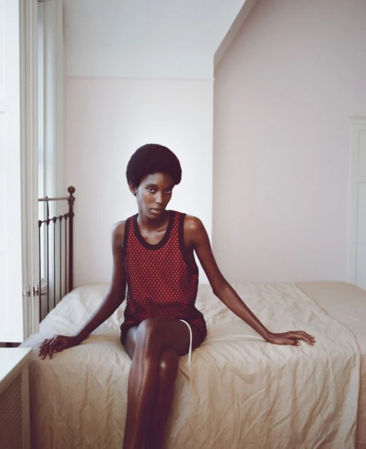 An adult woman with dark skin and a short afro hairstyle sits on a bed in a red mesh tank top and dark shorts in a bedroom.