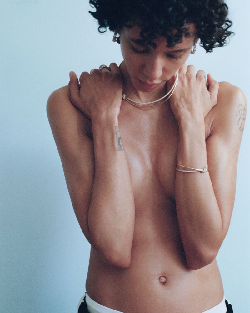 Close-up portrait of a slender, light-skinned woman with curly hair, accessorized with a thin necklace and bracelet, posing indoors.