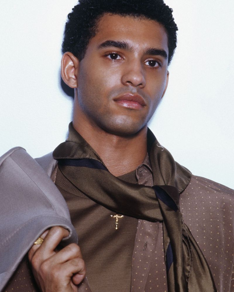 Close-up portrait of a stylish adult man with dark hair wearing a brown patterned shirt, scarf, and suit jacket in a photo studio.