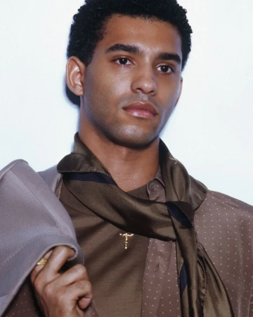Close-up portrait of a stylish adult man with dark hair wearing a brown patterned shirt, scarf, and suit jacket in a photo studio.