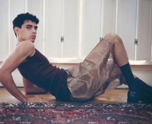 Model wearing a brown tank top and shiny shorts reclining on a wooden floor in front of white paneled doors.